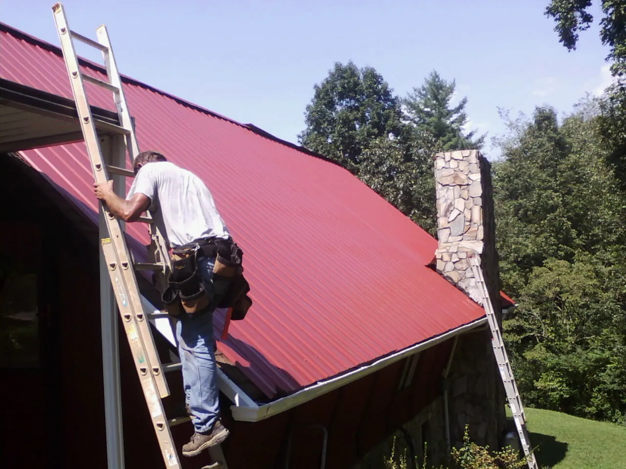 Roofer on a ladder beside a red metal roof and stone chimney — Soffit & Fascia Repair by Roofing Brothers in Fordyce